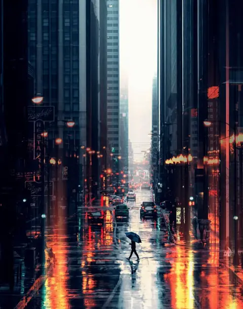 A person with an umbrella crosses a rainy Chicago street full of city light reflections