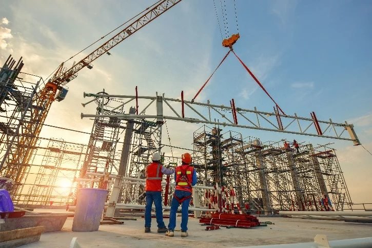 Two workers watching a tower crane lift a structural steel truss on a commercial jobsite