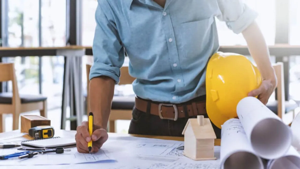 Contractor holding a hard hat and marking up blueprints next to a small model house