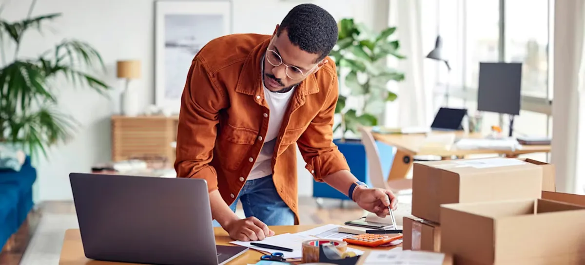 Online store owner packing orders and checking sales on a laptop