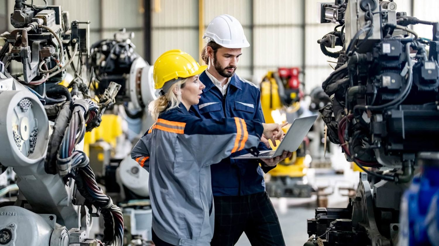 Two technicians reviewing a laptop between rows of industrial robotic arms