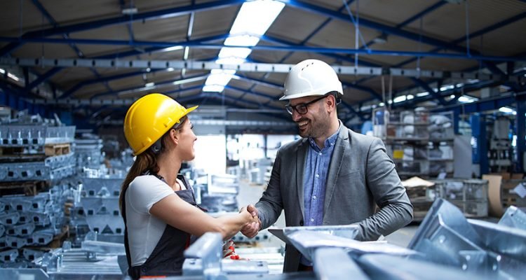 Plant manager and a buyer shaking hands on a factory floor beside stacked steel