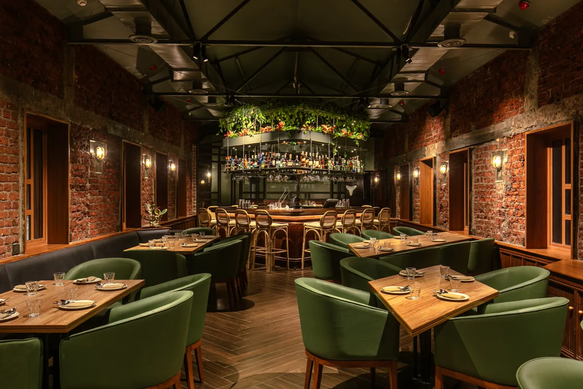 Restaurant dining room with green leather chairs, brick walls, and a lit bar