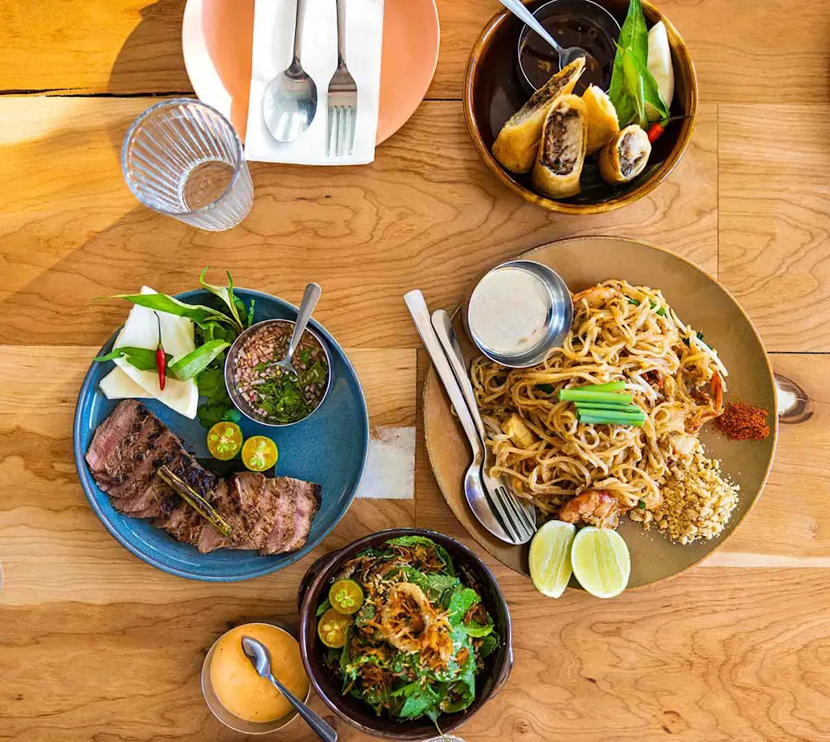 Overhead view of Thai dishes including pad thai, grilled beef, spring rolls, and salad