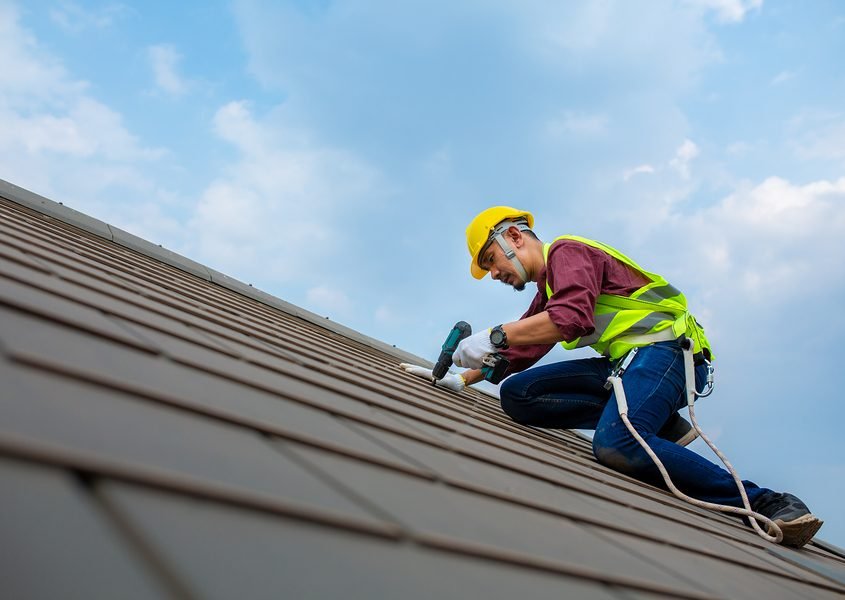 Roofer in a hard hat and safety harness fastening roof panels against a blue sky