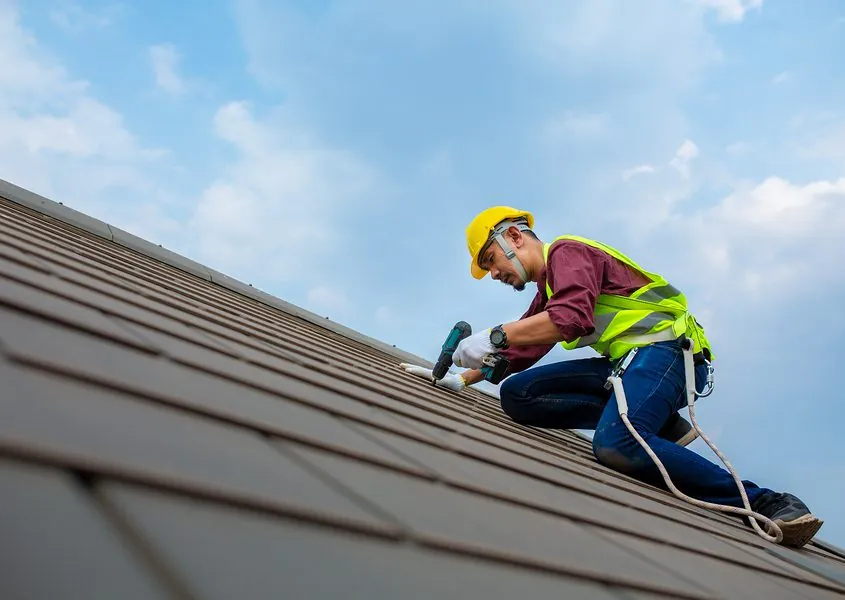 Roofer in a hard hat and safety harness fastening roof panels against a blue sky